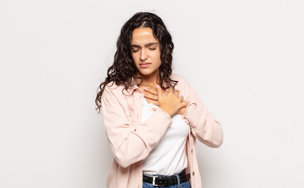 Foto mulher jovem e bonita parecendo triste, magoada e com o coração partido, segurando as duas mãos perto do coração, chorando e se sentindo deprimida