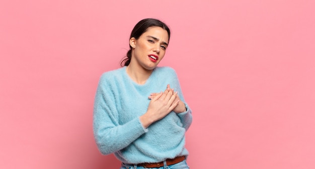 Mulher jovem e bonita parecendo triste, magoada e com o coração partido, segurando as duas mãos perto do coração, chorando e se sentindo triste