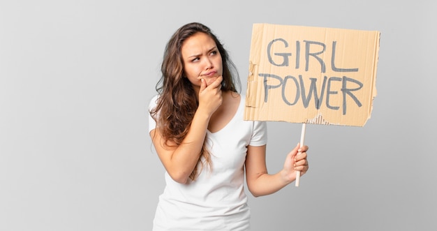 Mulher jovem e bonita pensando, sentindo-se em dúvida e confusa e segurando uma bandeira do poder feminino