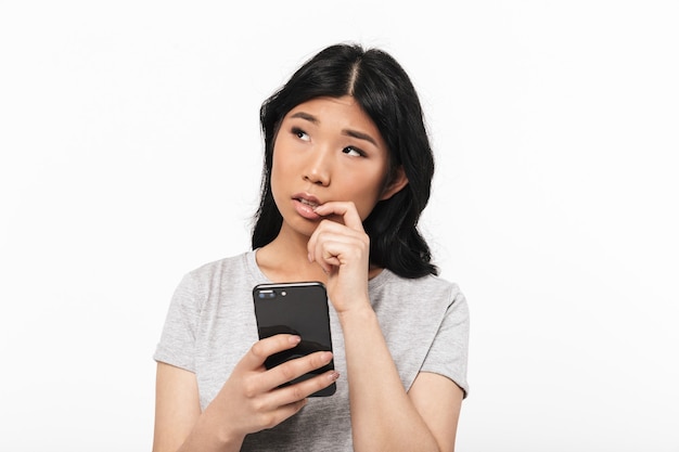 mulher jovem e bonita pensativa asiática posando isolado sobre uma parede branca, usando telefone celular.