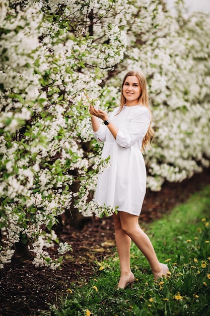 Mulher jovem e bonita perto de árvore branca florescendo no parque primavera. Uma mulher de vestido branco está entre as flores de uma macieira. conceito de temporada de primavera