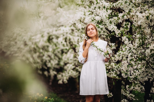 Mulher jovem e bonita perto de árvore branca florescendo no parque primavera. Uma mulher de vestido branco está entre as flores de uma macieira. conceito de temporada de primavera