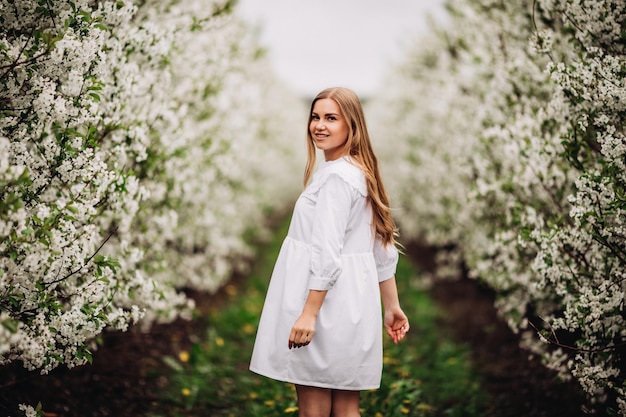 Mulher jovem e bonita perto de árvore branca florescendo no parque primavera. Uma mulher de vestido branco está entre as flores de uma macieira. conceito de temporada de primavera