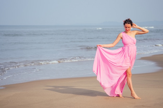 Mulher jovem e bonita posando em um vestido rosa longo chique de luxo elegante na praia com ondas. conceito de férias chiques em um resort tropical.