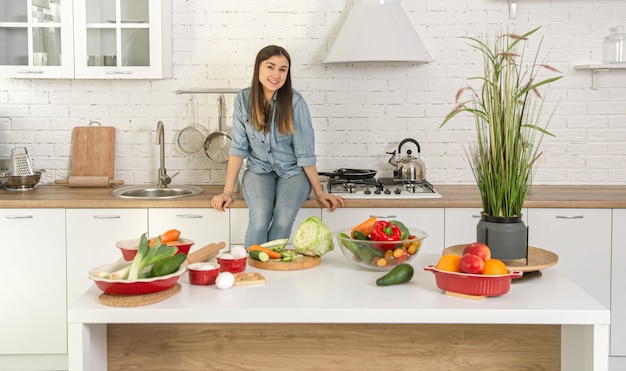 Mulher jovem e bonita preparando uma salada vegetariana de vários vegetais no fundo do interior da cozinha em cores claras.