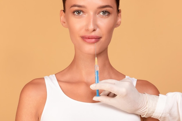 Mulher jovem e bonita recebendo injeção facial sobre cosmetologista de fundo bege segurando seringa perto do queixo