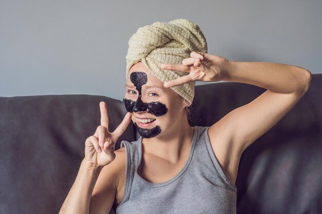 Mulher jovem e bonita relaxando com máscara facial em casa. Mulher feliz e alegre aplicando máscara preta no rosto