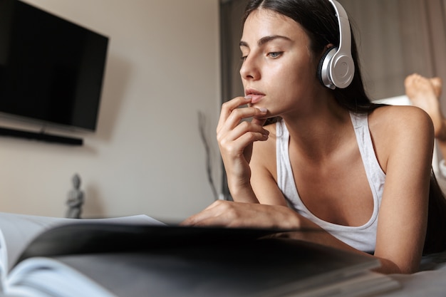 Mulher jovem e bonita relaxando em um sofá em casa, ouvindo música com fones de ouvido, lendo um livro