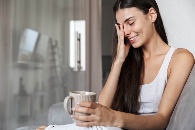 Mulher jovem e bonita relaxando em um sofá em casa, segurando a xícara