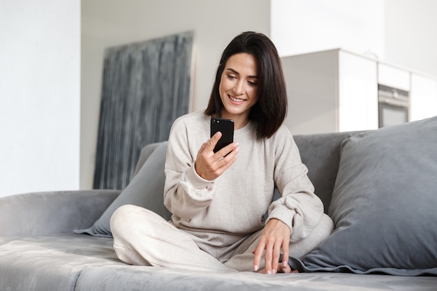 Mulher jovem e bonita relaxando em um sofá em casa, usando o telefone celular