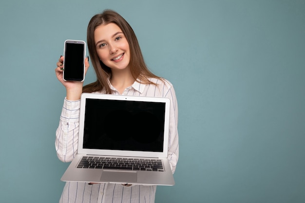 Mulher jovem e bonita satisfeita sorridente segurando computador, laptop e celular, olhando para a câmera