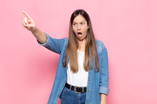 Mulher jovem e bonita se sentindo chocada e surpresa, apontando e olhando para cima com admiração e expressão boquiaberta