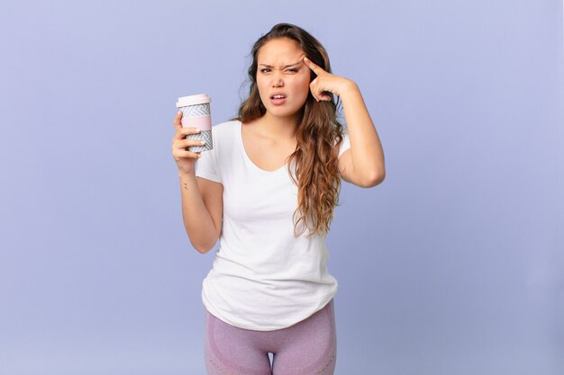 Mulher jovem e bonita se sentindo confusa e perplexa, mostrando que você é maluca e segurando um café