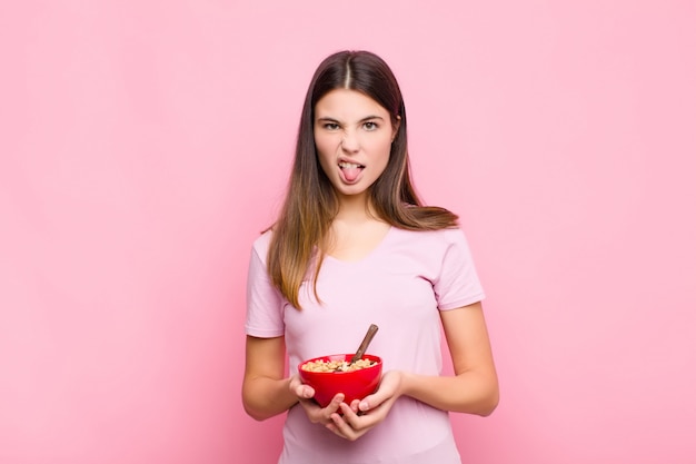 Mulher jovem e bonita se sentindo enojada e irritada, enfiando a língua para fora, não gostando de algo desagradável e nojento com uma tigela de café da manhã