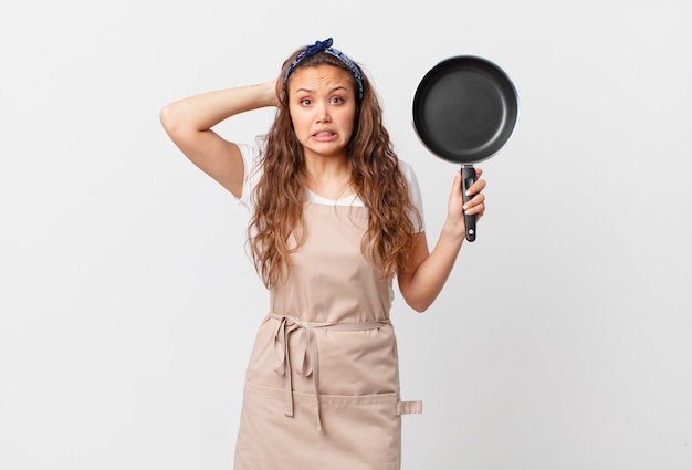 Mulher jovem e bonita se sentindo estressada, ansiosa ou com medo, com as mãos no conceito do chef principal e segurando uma panela