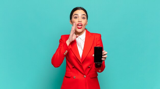 Mulher jovem e bonita se sentindo feliz, animada e positiva, dando um grande grito com as mãos perto da boca, gritando. conceito de telefone inteligente