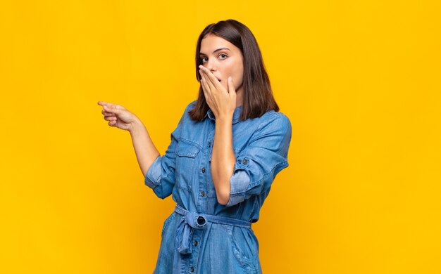 Mulher jovem e bonita se sentindo feliz, chocada e surpresa, cobrindo a boca com a mão e apontando para o espaço lateral da cópia