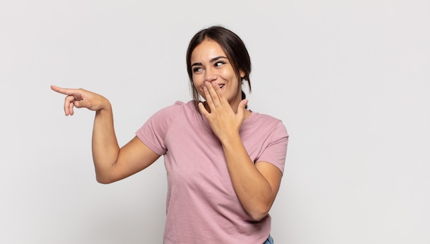 Mulher jovem e bonita se sentindo feliz, chocada e surpresa, cobrindo a boca com a mão e apontando para o espaço lateral da cópia