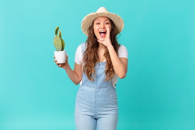 Mulher jovem e bonita se sentindo feliz, dando um grande grito com as mãos perto da boca e segurando um cacto