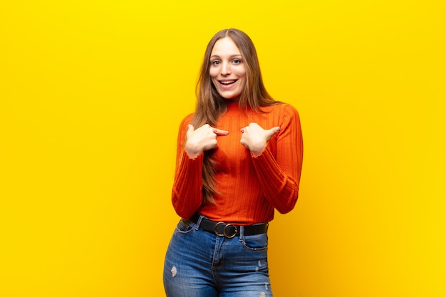 Mulher jovem e bonita se sentindo feliz, surpresa e orgulhosa, apontando para si mesma com um olhar excitado e espantado contra a parede laranja