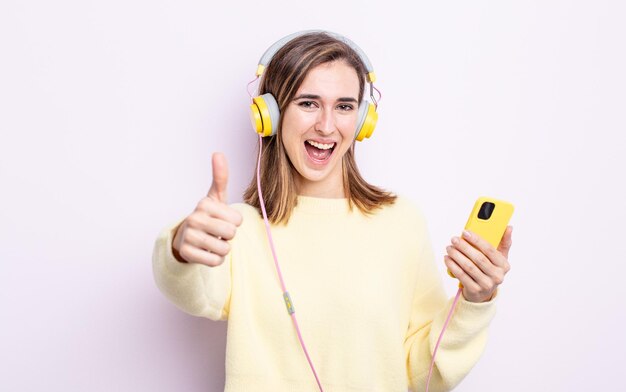 Mulher jovem e bonita se sentindo orgulhosa, sorrindo positivamente com os polegares para cima. fones de ouvido e conceito de telefone