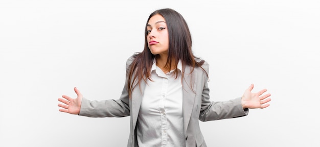 Mulher jovem e bonita se sentindo sem noção e confusa, sem ter ideia, absolutamente intrigada com um olhar idiota ou tolo isolado contra a parede branca