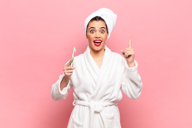 Mulher jovem e bonita se sentindo um gênio feliz e animado depois de realizar uma ideia, levantando o dedo alegremente, eureka !. conceito de roupão e escova de dentes