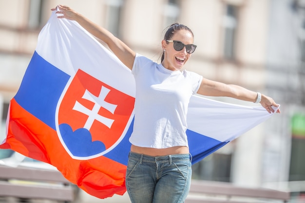 Mulher jovem e bonita segura uma bandeira da Eslováquia oudoors em um dia ensolarado, usando óculos escuros.