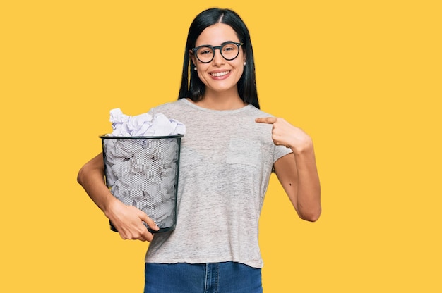 Mulher jovem e bonita segurando a caixa de papel cheia de papéis amassados apontando o dedo para si mesma sorrindo feliz e orgulhoso