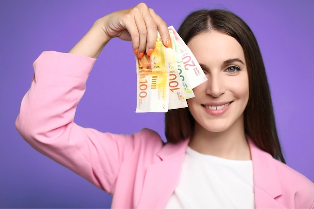 Mulher jovem e bonita segurando notas de shekels de israel perto do rosto sorrindo feliz com a boca aberta