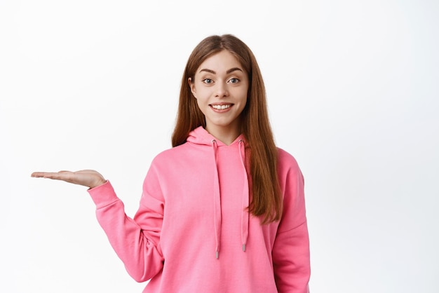 Mulher jovem e bonita segurando o produto exibe sua promoção na palma da mão contra o espaço branco da cópia sorrindo para a câmera em pé sobre fundo branco