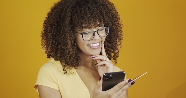 Mulher jovem e bonita segurando o smartphone e o cartão do banco de crédito. transação de compras online. método sem dinheiro de banco móvel. jovem sorridente afro-americana no fundo da parede amarelo-laranja