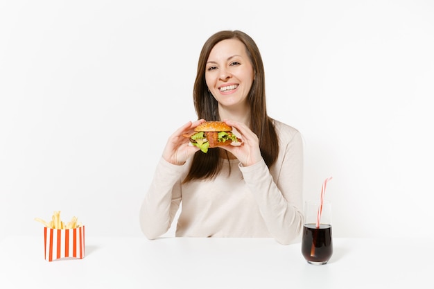 Foto mulher jovem e bonita sentada à mesa com hambúrguer, batata frita, coca-cola em frasco de vidro isolado no fundo branco. nutrição adequada ou fast food clássico americano. área de publicidade com espaço de cópia.