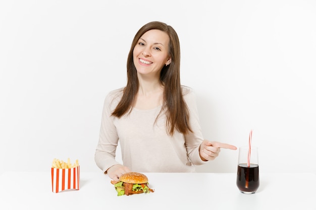 Mulher jovem e bonita sentada à mesa com hambúrguer, batata frita, coca-cola em frasco de vidro isolado no fundo branco. Nutrição adequada ou fast food clássico americano. Área de publicidade com espaço de cópia.
