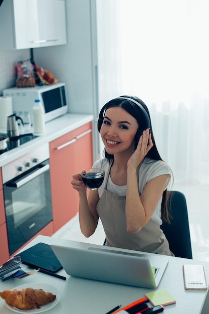 Mulher jovem e bonita sentada à mesa da cozinha com um laptop e segurando uma xícara de café enquanto toca seus fones de ouvido sem fio