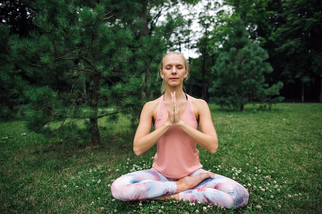 Foto mulher jovem e bonita sentada em pose de lótus de ioga no parque e meditando na grama verde