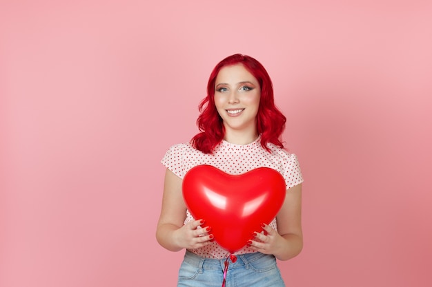 Mulher jovem e bonita sorridente com cabelo vermelho segurando um grande balão vermelho voador em forma de coração