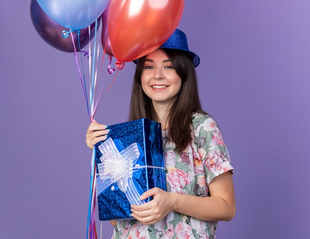 Mulher jovem e bonita sorridente com chapéu de festa segurando balões com uma caixa de presente isolada na parede azul