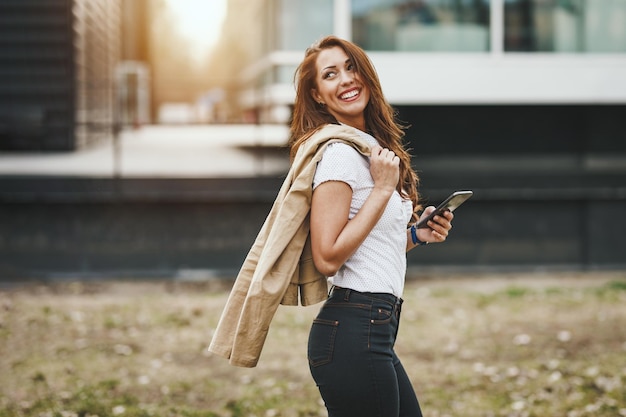Mulher jovem e bonita sorridente com smartphone na mão contra o fundo urbano da cidade.