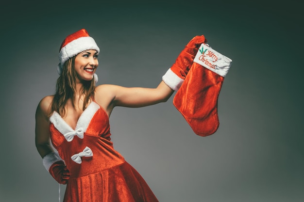 Mulher jovem e bonita sorridente em fantasia de Papai Noel pensando e segurando a meia vermelha de Natal.