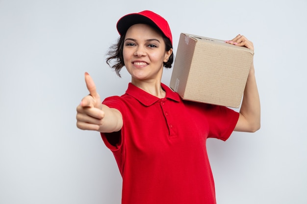 Mulher jovem e bonita sorridente, entregadora, segurando uma caixa de papelão no ombro e apontando para a frente