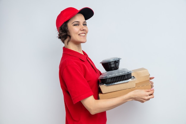 Mulher jovem e bonita sorridente, entregando-se de pé, segurando recipientes de comida e embalagens em caixas de pizza