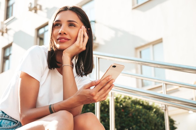 Mulher jovem e bonita sorridente hipster em roupas da moda de verão Mulher despreocupada sexy posando no fundo da rua ao pôr do sol Modelo positivo usando aplicativos de smartphone Segurando o telefone olhando para o celular