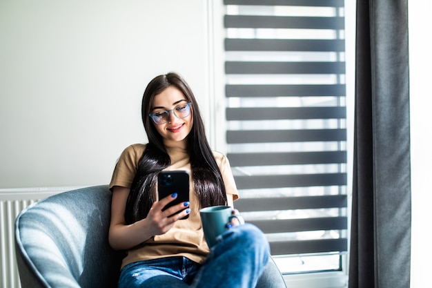 Mulher jovem e bonita sorridente olhando para seu telefone inteligente enquanto está sentado em uma cadeira grande e confortável em casa