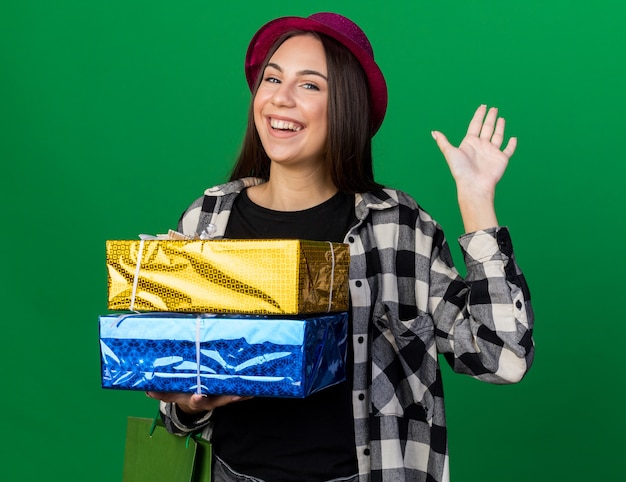 Mulher jovem e bonita sorridente usando um chapéu de festa segurando uma caixa de presente e uma sacola de presentes mostrando um gesto de olá isolado na parede verde