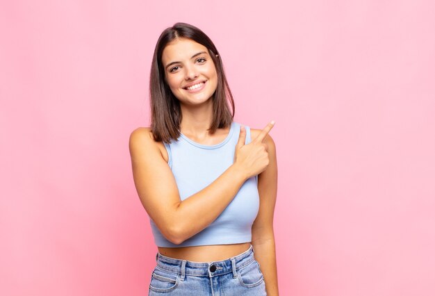 Mulher jovem e bonita sorrindo alegremente, sentindo-se feliz e apontando para o lado e para cima, mostrando o objeto no espaço da cópia