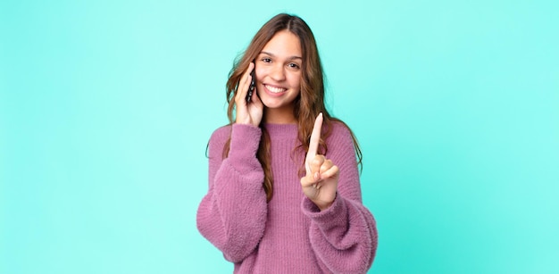 Mulher jovem e bonita sorrindo com orgulho e confiança, alcançando o primeiro lugar e usando um smartphone