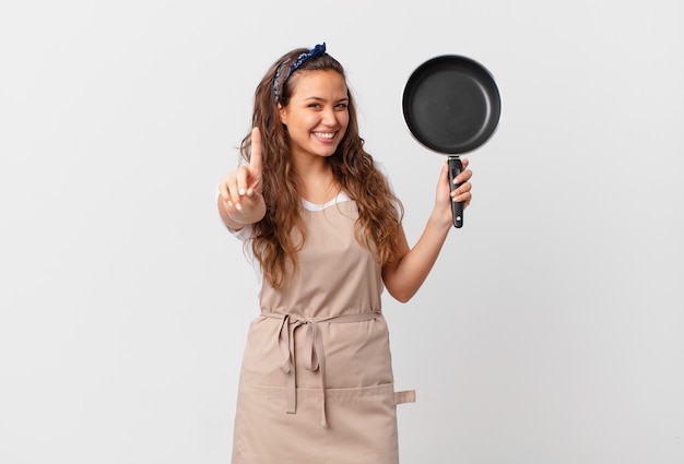 Mulher jovem e bonita sorrindo com orgulho e confiança, fazendo o conceito de chef número um e segurando uma panela