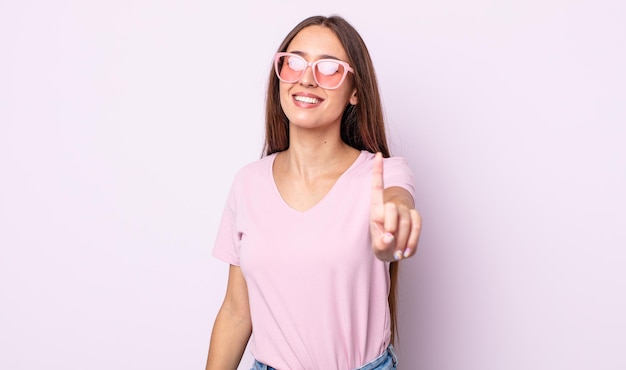 Mulher jovem e bonita sorrindo com orgulho e confiança, fazendo o número um. conceito de óculos de sol rosa