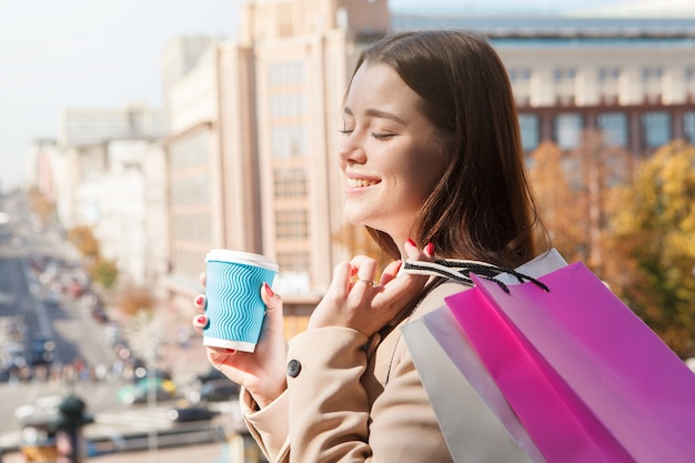 Mulher jovem e bonita sorrindo com os olhos fechados, tomando um café ao sol em um dia quente de outono, depois de fazer compras na cidade